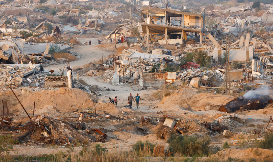 A devastated landscape of collapsed buildings and rubble, with people walking through the ruins after widespread destruction.